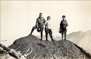 One man and two women in hiking clothes and boots stand on a rock with mountain peaks in the background